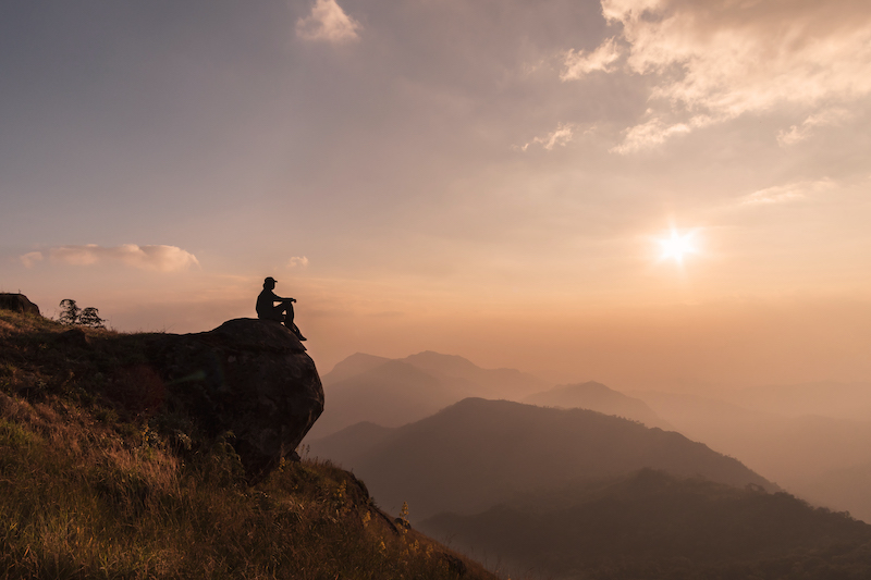 Un étudiant de l'Institut Atlas assis paisiblement sur un rocher au sommet d'une montagne, contemplant le lever du soleil au-dessus d'une chaîne de montagnes brumeuses, capturant l'essence de l'apprentissage et de l'exploration en tourisme.
